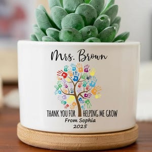 Könnte beinhalten: Weißer Keramik-Pflanztopf mit einer Sukkulente. Der Topf hat ein farbenfrohes Handabdruck-Baum-Design und den Text "Mrs. Brown, THANK YOU FOR HELPING ME GROW From Sophia 2025". Er steht auf einem runden Holzuntersetzer.