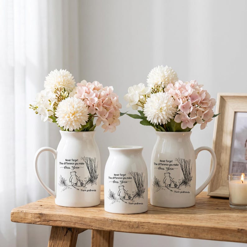 May include: Three white ceramic vases with handles and a cylindrical vase, each with a black and white illustration of a bear and text. The vases hold bouquets of white and pink flowers. The text reads "Never forget the difference you make Mrs. Gloria. Thank you friends."