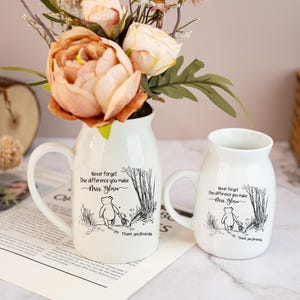 May include: Two white ceramic mugs with handles, one tall and one short, each featuring a Winnie the Pooh illustration and the text "Never forget the difference you make Mrs. Gloria. Thank you, Brenda.". The mugs are filled with flowers.