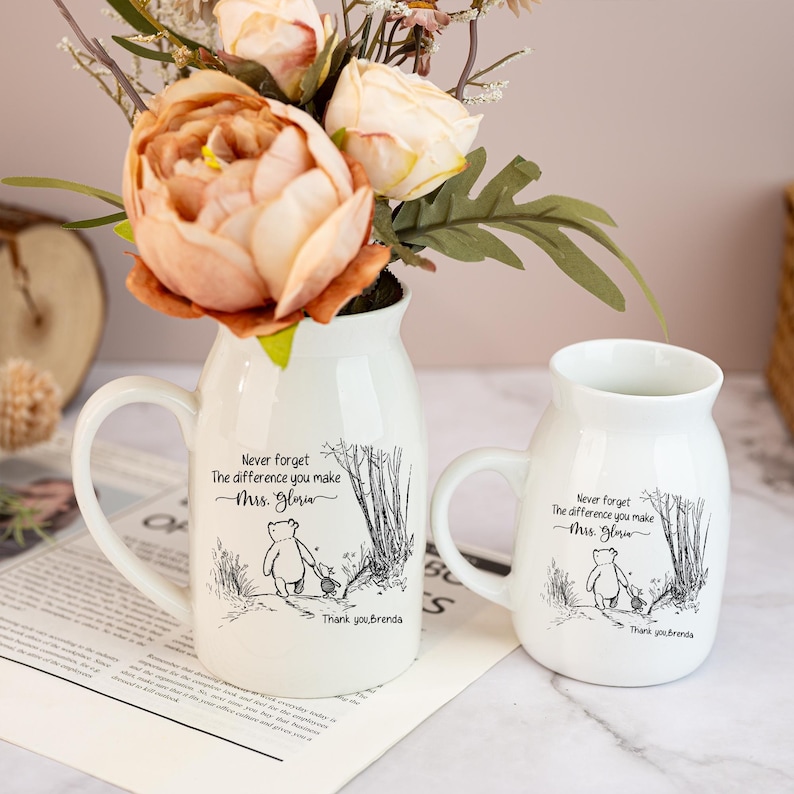 May include: Two white ceramic mugs with handles, one tall and one short, each featuring a Winnie the Pooh illustration and the text "Never forget the difference you make Mrs. Gloria. Thank you, Brenda.". The mugs are filled with flowers.