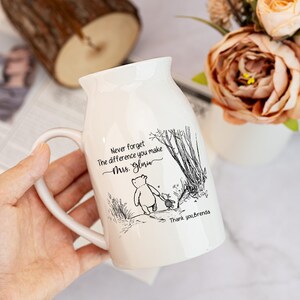 May include: White ceramic mug with a curved handle, featuring a black and white illustration of a bear and a small character walking in a forest scene. The mug has the text "Never forget the difference you make Mrs. Gloria" and "Thank you, brenda".