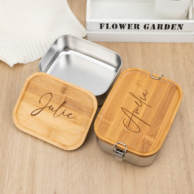 May include: Two stainless steel lunch boxes with bamboo lids. The lids are engraved with the names "Julie" and "Amelia".