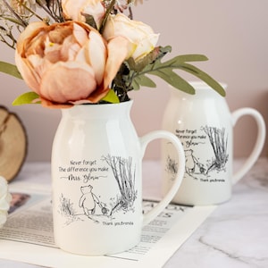 May include: White ceramic vase with a handle, featuring a Winnie the Pooh illustration and the text "Never forget the difference you make Mrs. Gloria. Thank you, Brenda." The vase holds peach and cream-colored flowers, perfect for home decor.