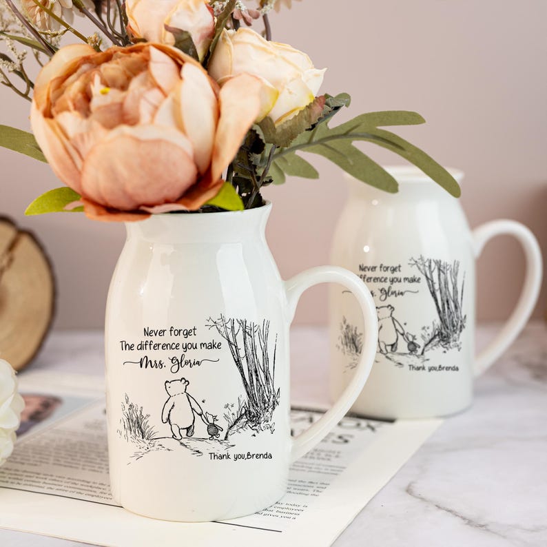 May include: White ceramic vase with a handle, featuring a Winnie the Pooh illustration and the text "Never forget the difference you make Mrs. Gloria. Thank you, Brenda." The vase holds peach and cream-colored flowers, perfect for home decor.