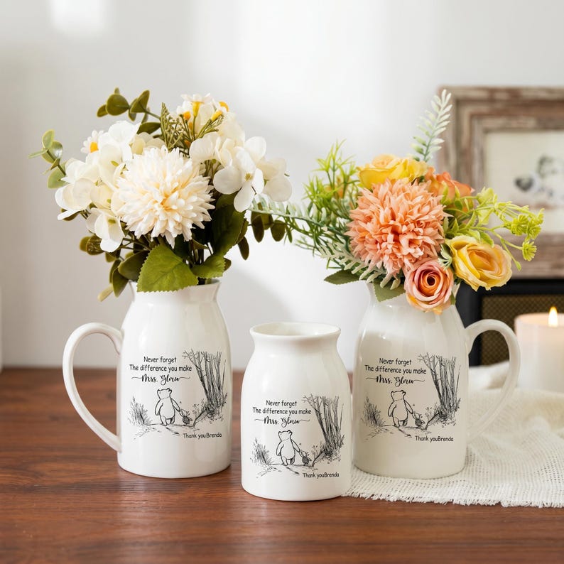 May include: Three white ceramic vases with floral arrangements. The vases have a handle and are decorated with a black and white illustration of a bear and text that reads "Never forget the difference you make Mrs. [Name] Thank you [Name]"