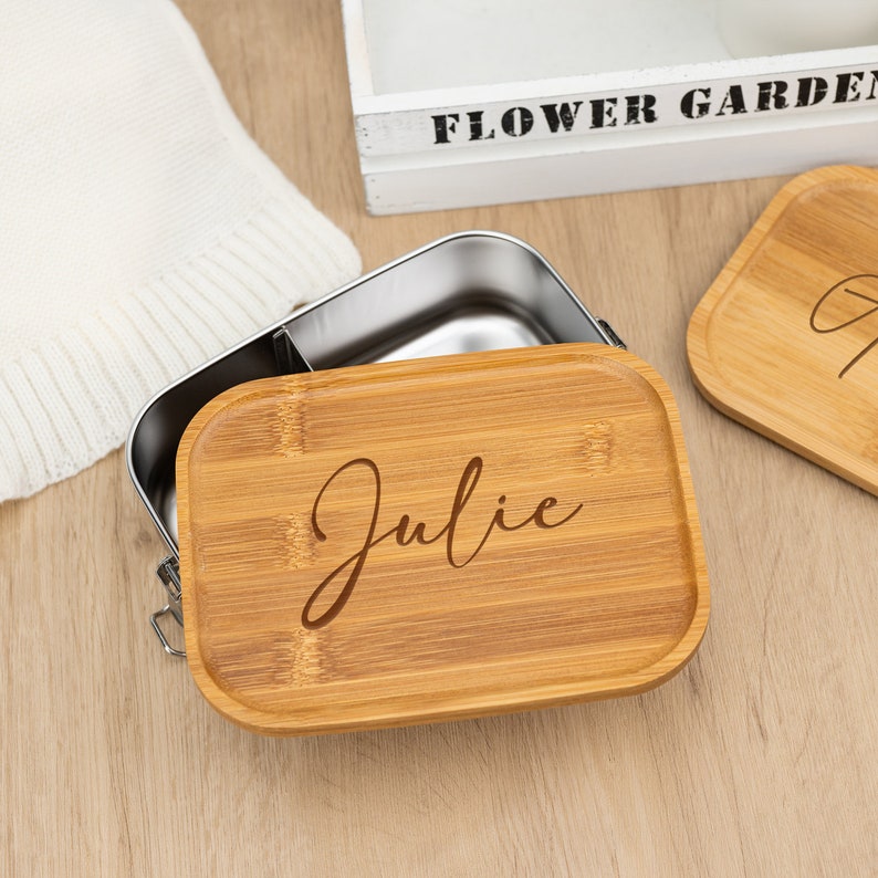 May include: A stainless steel lunch box with a bamboo lid. The lid is engraved with the name "Julie".