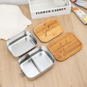 May include: Two stainless steel lunch boxes with a divider in one of the boxes. The lunch boxes have wooden lids with the names "Julie" and "Amelia" engraved on them.