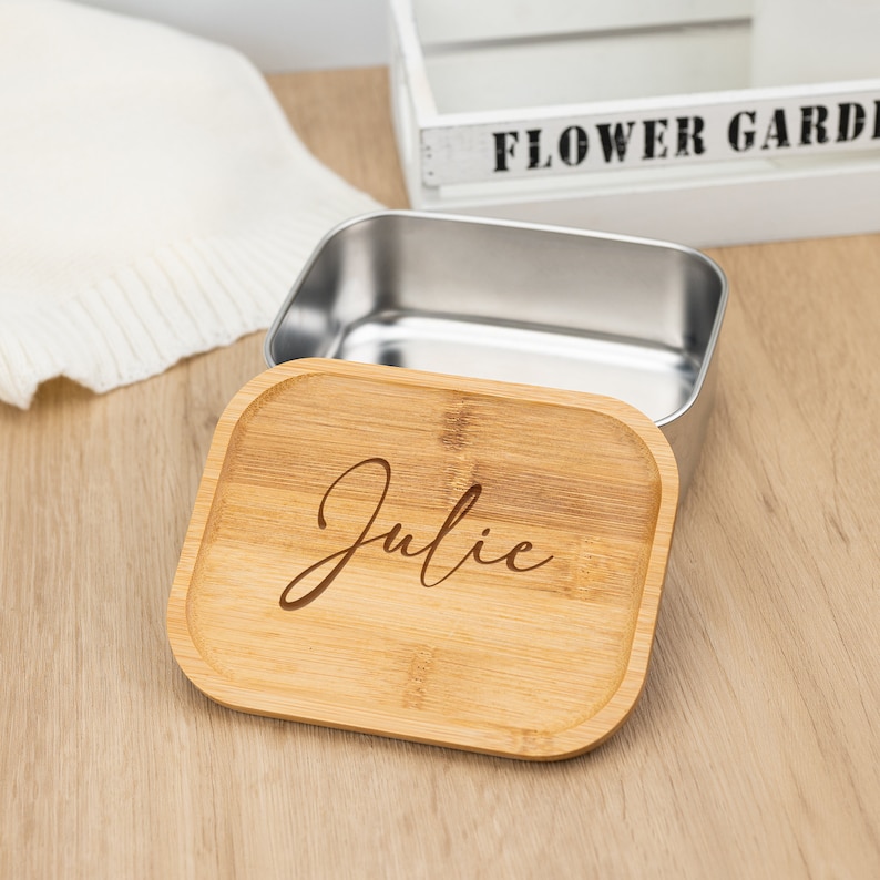 May include: A wooden lunchbox with a stainless steel interior. The lid is made of bamboo and has the name "Julie" engraved on it.