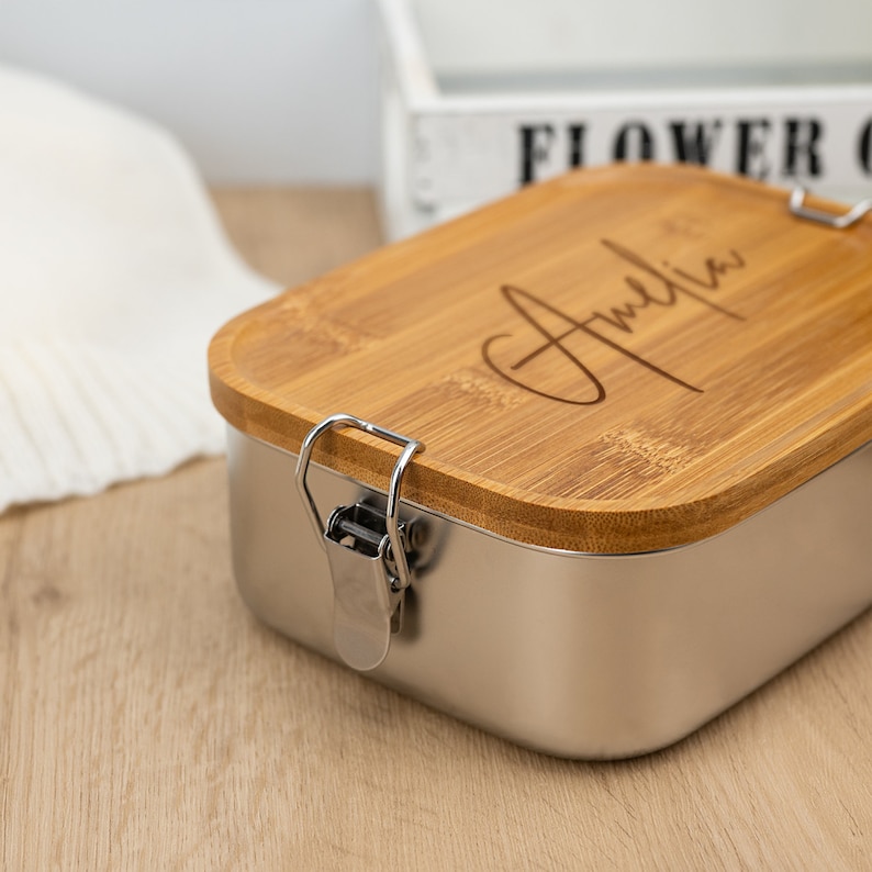 May include: A stainless steel lunch box with a bamboo lid. The lid has a latch and is engraved with the name "Amelia".