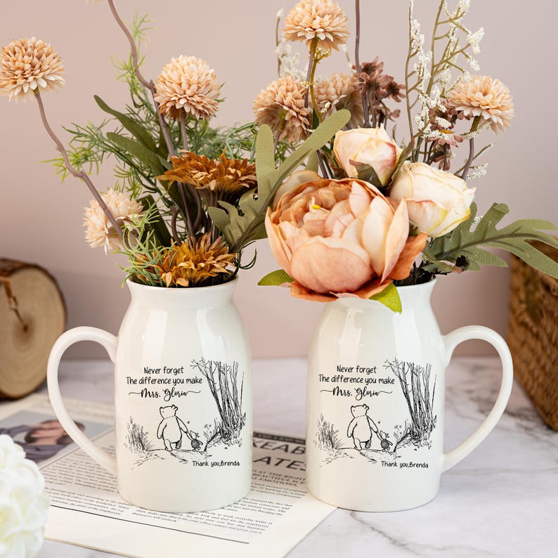 May include: Two white ceramic vases with handles, each holding faux flowers in shades of peach, cream, and brown. The vases have a black and white illustration and text that reads "Never forget the difference you make Mrs. Gloria. Thank you, Brenda."
