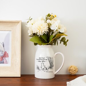 May include: A white ceramic vase with a handle, filled with white and yellow flowers and green leaves. The vase has a black illustration of a bear and text that reads "Never forget the difference you make". A framed picture is to the left.