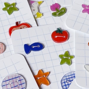 Peut inclure: Une collection de boutons en plastique translucide coloré aux formes variées, notamment des étoiles, un poisson, une tomate, une fleur et des motifs circulaires, fixés sur des cartes de présentation individuelles à quadrillage blanc.