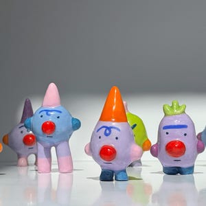 Op de afbeelding: Een verzameling grillige keramische beeldjes in diverse kleuren. Elk figuur heeft een uniek ontwerp, met een rode neus, geschilderde ogen en een kegelvormige of kroonvormige kop. De beeldjes worden tentoongesteld op een wit oppervlak.