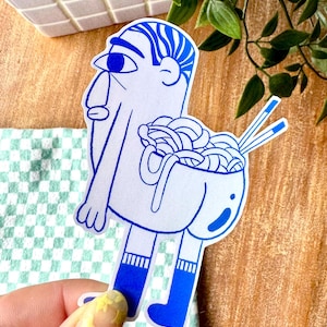 Peut inclure: Un autocollant blanc représentant un personnage de dessin animé bleu avec un bol de nouilles pour le torse. Le personnage a des cheveux bleus, un grand œil et porte des chaussettes et des bottes bleues. Des baguettes sont placées sur le côté droit du bol.