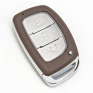 May include: A car key fob with a brown textured casing and a silver metallic centre panel. The panel features three buttons with icons for locking, unlocking, and opening the boot. A silver metallic loop is located at the bottom of the device.