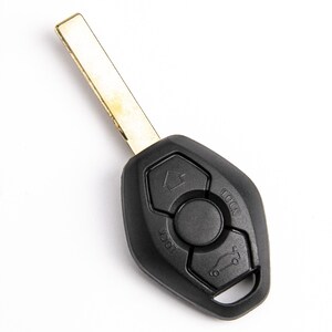 May include: A black plastic car key fob with a metallic blade. The fob features three buttons: one with an upward arrow icon, one with a car trunk icon, and a central circular button. The word LOCK is embossed twice near the button icons.