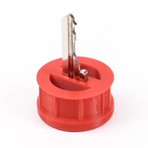 Könnte beinhalten: Ein roter, 3D-gedruckter Schlüsselhalter mit einem silbernen Schlüssel. Der Schlüsselhalter hat ein kreisförmiges Design mit einem Schlitz, in dem der Schlüssel aufrecht stehen kann. Der Schlüssel ist aus Metall und hat ein abgenutztes Aussehen.