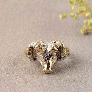 Może przedstawiać: Złoty pierścionek z detalicznym wzorem głowy barana. Rogi barana są zakrzywione, a głowa jest fakturowana.