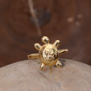 May include: A gold-colored ring with a sun design. The sun has a face with a small nose and two eyes.
