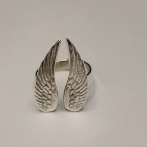 Puede incluir: Un anillo de plata con un diseño de dos alas de ángel.