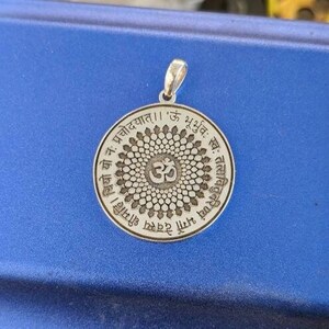 May include: A silver pendant with a circular design featuring a mandala with a stylized Om symbol in the centre. The pendant is inscribed with Sanskrit text, including the phrase 'Om Bhu Bhuvah Suvah'.