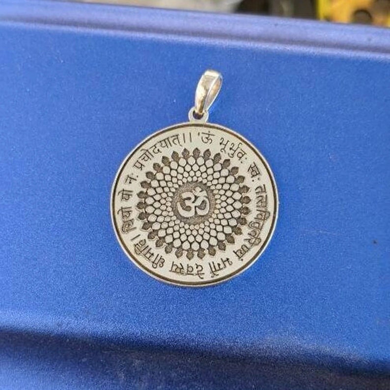 May include: A silver pendant with a circular design featuring a mandala with a stylized Om symbol in the centre. The pendant is inscribed with Sanskrit text, including the phrase 'Om Bhu Bhuvah Suvah'.