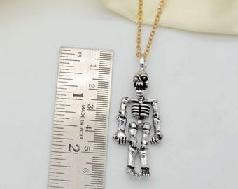 May include: Silver skeleton pendant necklace with a gold chain. The pendant is about 2 inches tall and is on a white surface next to a ruler.