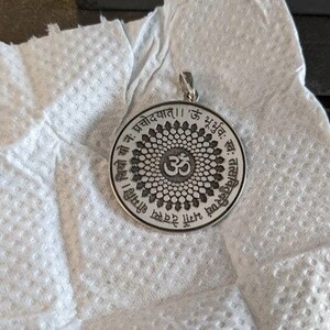 May include: Silver pendant with a circular design featuring an Om symbol surrounded by a mandala pattern. The pendant is inscribed with Sanskrit text: "Om Gam Ganapataye Namaha" and "Om Shri Maha Ganapataye Namaha".