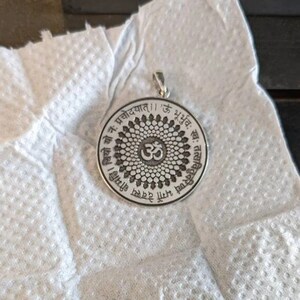 May include: A silver pendant with a circular design featuring the Om symbol surrounded by a mandala and Sanskrit text. The text reads "Om Shanti Shanti Shanti" and "Om Gam Ganapataye Namaha".