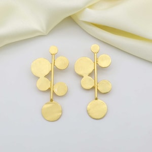 May include: A pair of gold-coloured, abstract drop earrings. Each earring has a vertical stem with circular and semi-circular shapes. The earrings have a matte finish and are displayed on a white surface with a cream-coloured fabric in the background.