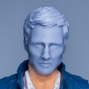 May include: A blue action figure head with short, styled hair. The figure is wearing a blue jacket over a white collared shirt. The skin tone is a pale blue color. The figure is set against a light blue background.