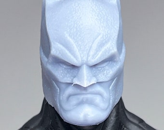 May include: A close-up of a Batman action figure head sculpt. The head is made of a light gray plastic and has a detailed sculpt of Batman's face. The head is attached to a black cape and a black and gray suit.