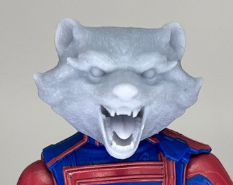 May include: A blue and red action figure with a gray raccoon head. The figure is wearing a black belt and has a gold emblem on the chest.