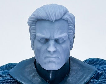 May include: A close-up of a gray plastic action figure head with a serious expression. The figure is wearing a blue cape and a black and gray chest piece with a bat symbol.