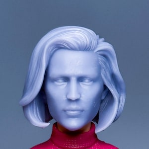 Op de afbeelding: Een plastic pop met kort, lichtblauw haar, die een roze trui draagt. De pop heeft een neutrale gezichtsuitdrukking.