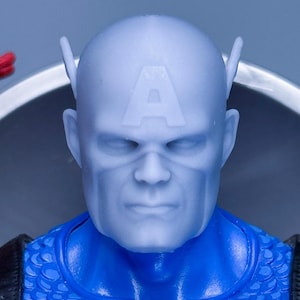 Puede incluir: Primer plano de una figura de acción del Capitán América. La figura tiene una estrella azul y blanca en el pecho, un traje azul con estampado de escamas y una "A" blanca en la frente. Expresión seria, correas negras.