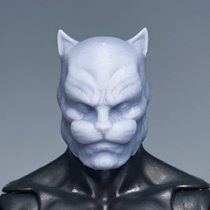 May include: A close-up of a gray, unpainted plastic action figure head with a cat-like face. The head has pointed ears, a wide nose, and a small mouth. The figure is wearing a black suit.