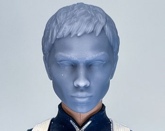 May include: A close-up of a female action figure's head and torso. The figure has short, gray hair and is wearing a dark blue jumpsuit with a white zipper and a white strap across the chest.