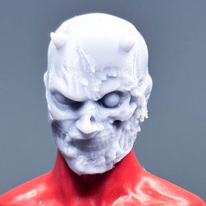 Könnte beinhalten: Nahaufnahme einer weißen, strukturierten Maske mit schädelartigem Aussehen, kleinen Hörnern und einem bedrohlichen Ausdruck. Die Maske ist vor einem roten Torso platziert, was einen auffälligen Kontrast erzeugt. Es ist eine Sammler-Actionfigur.