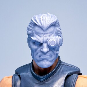 May include: A gray plastic action figure head with a detailed face and a small, round, black eye piece on the right side.