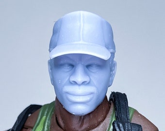 May include: A close-up of a dark-skinned action figure wearing a camouflage tank top and a light blue baseball cap. The figure is wearing a black harness with two black pouches and two green pouches.