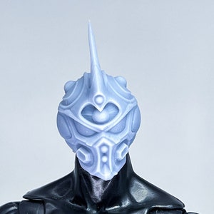 Puede incluir: Un casco impreso en 3D, de color azul claro, detallado con una parte superior puntiaguda y un diseño circular. El casco tiene una gran abertura central para los ojos y aberturas circulares más pequeñas alrededor de los bordes.