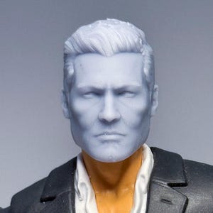 Op de afbeelding: Een lichtblauwe actiefiguurkop met kort, gestyled haar. De figuur draagt een wit overhemd met kraag en een zwarte jas. De figuur heeft een serieuze uitdrukking.