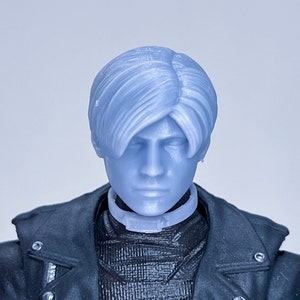 Peut inclure: Gros plan sur la tête d'une figurine en plastique avec des cheveux courts et foncés et une veste en cuir noire. La figurine a une expression neutre et porte un collier noir.