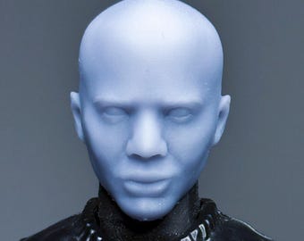 Puede incluir: Primer plano de una cabeza calva azul claro con rasgos faciales detallados, incluyendo ojos, nariz y labios. La cabeza está unida a una prenda negra texturizada con cuello alto. El fondo es gris liso.