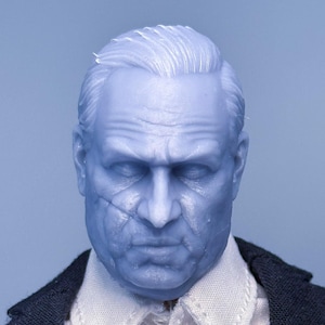 May include: A close-up of a man's face, with a white shirt and a dark blue suit jacket. The man has short, gray hair and a serious expression.