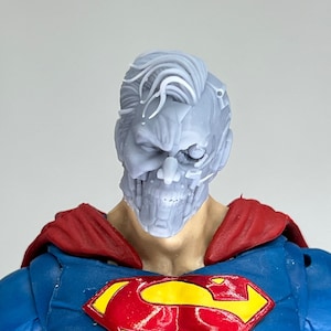 May include: A gray plastic action figure head with a detailed, damaged face and a Superman logo on the chest. The figure is wearing a red and blue costume.
