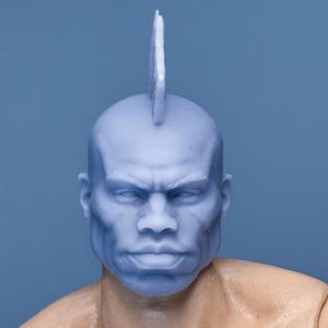 May include: A blue plastic action figure head with a mohawk hairstyle. The head is sculpted with a realistic face and a defined jawline.