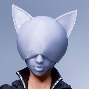 Puede incluir: Una máscara gris de Catwoman con orejas puntiagudas, que cubre la mitad superior de la cara de una figura. La figura lleva una chaqueta negra con cremallera y cuello en V. La máscara tiene un acabado liso y mate.