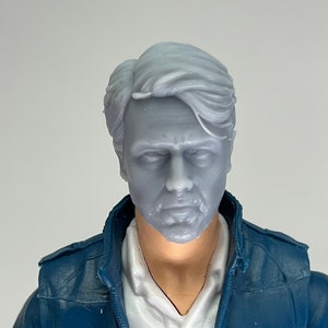 May include: A close-up of a man's head and shoulders wearing a blue jacket over a white shirt. The man has short, gray hair and a beard.